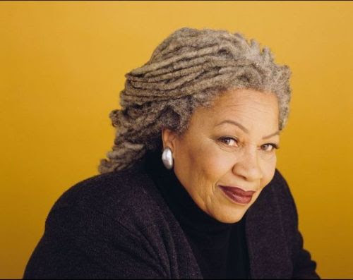 Siting against a mustard background is the author Toni Morrison, dressed in black, her hair in dreadlocks