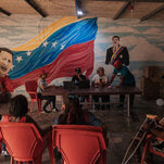 People sitting on a table in front of a mural depicting the Venezuelan flag and its president, Nicolás Maduro.