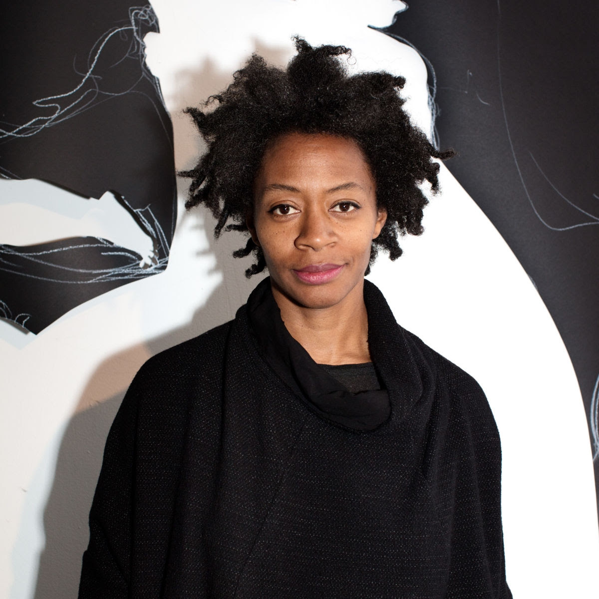 Artist Kara Walker stands in front of a black-and-white wall, wearing black and lightly smiling.