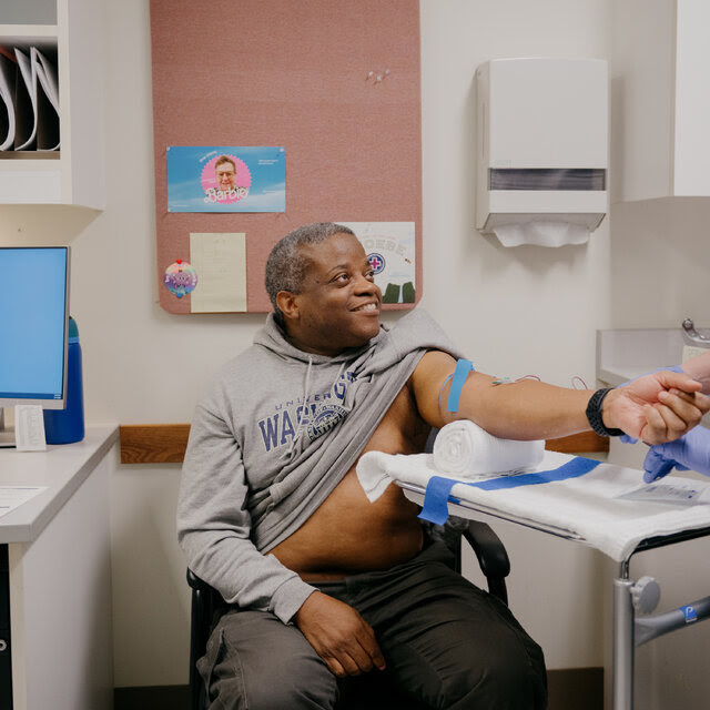 Kenneth Davis sits in a clinic with a gray sweatshirt lifted over his stomach and his arm extended to a standing health care worker, who is drawing blood.