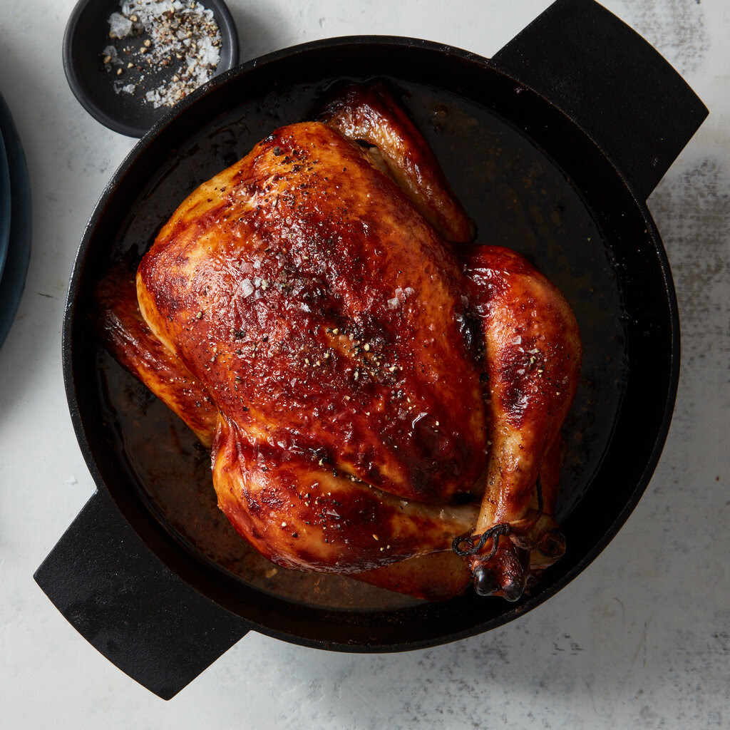 A deeply burnished buttermilk-brined roast chicken is shown in a cast-iron pot with a stack of blue plates and a small dish of salt and pepper nearby.