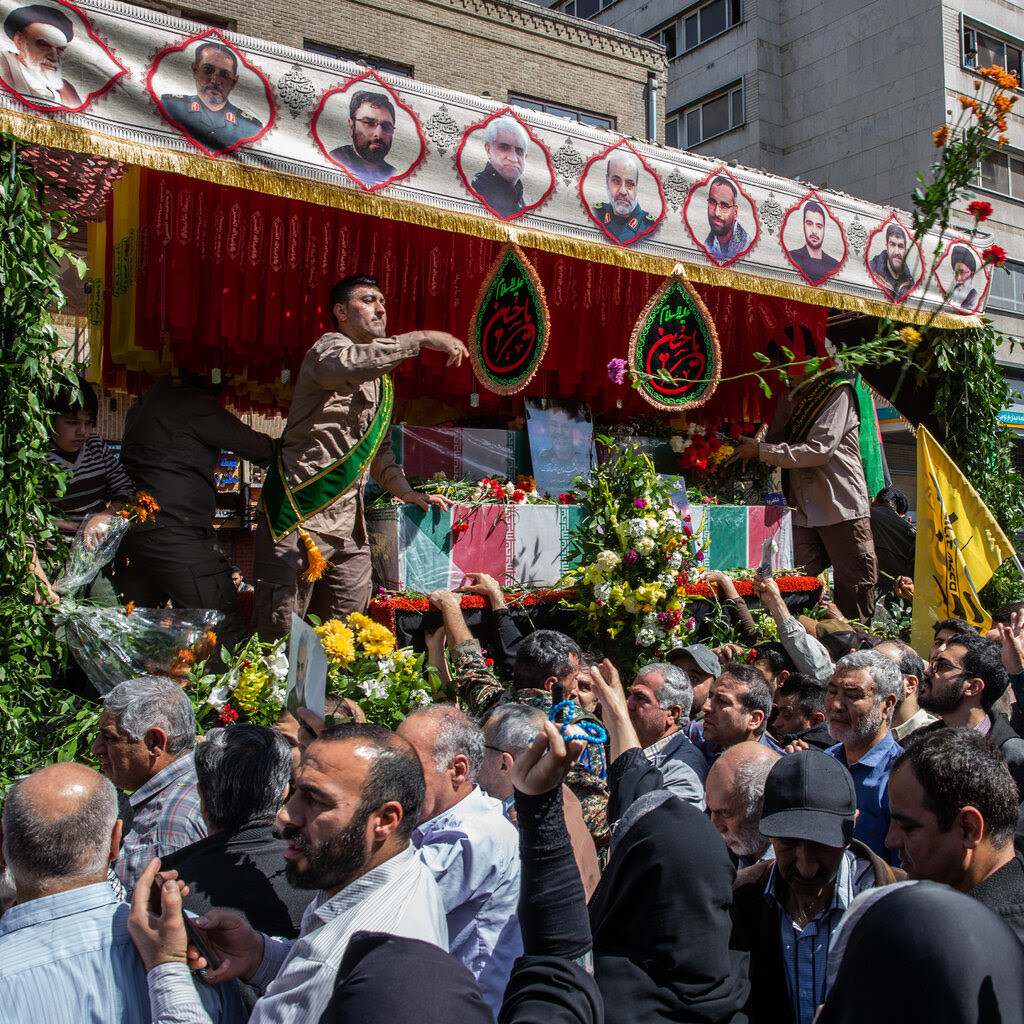 A man throws flowers from a stage lined with a banner showing several men’s faces as a large crowd passes by.