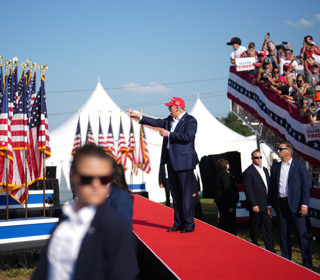 Mr. Trump pointing in front of him as he walks on a red carpet at an outdoor rally with a crowd behind him and security detail nearby.