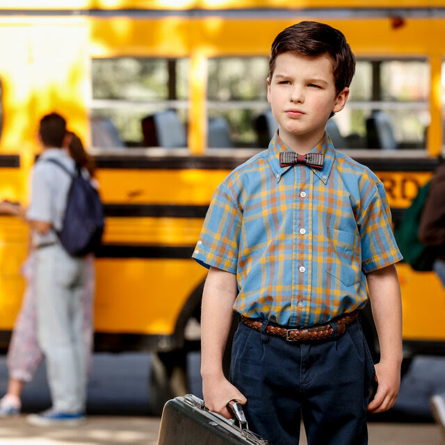 Iain Armitage as the young Sheldon Cooper stands in front of a yellow school bus holding a briefcase.