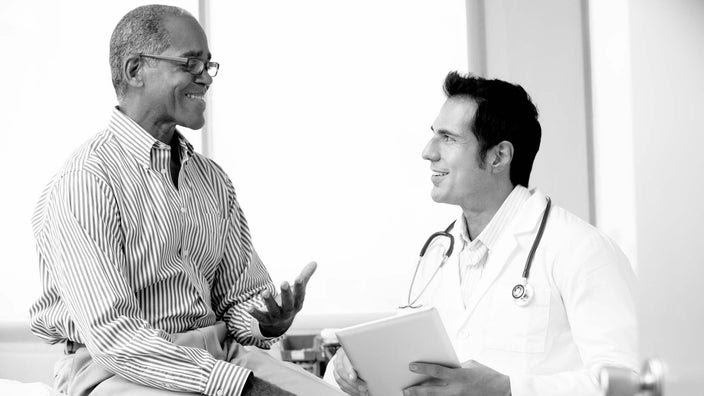 Black-and-white image of an older man consulting with his doctor in the exam room.