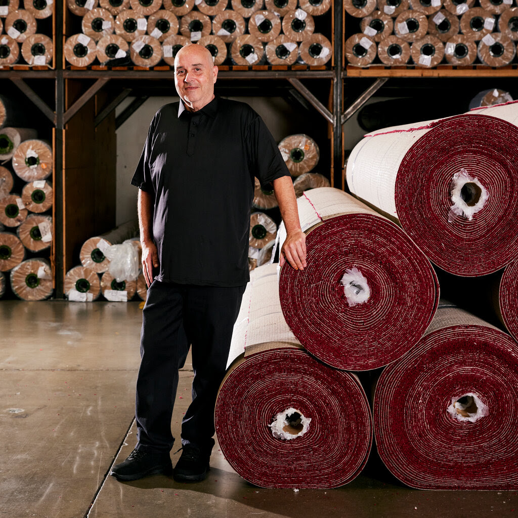 Steve Olive in a black shirt and black pants stands near a roll of red carpets in a warehouse.