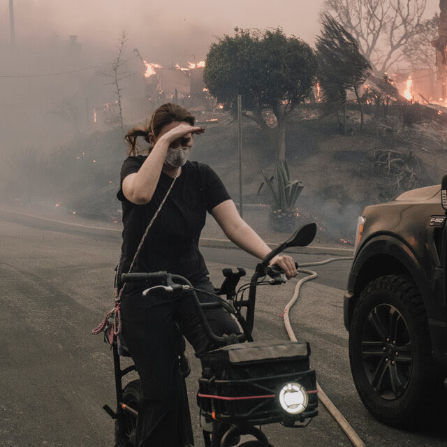 A woman shields her eyes as she rides a bike on a road blanketed by smoke from fires nearby.