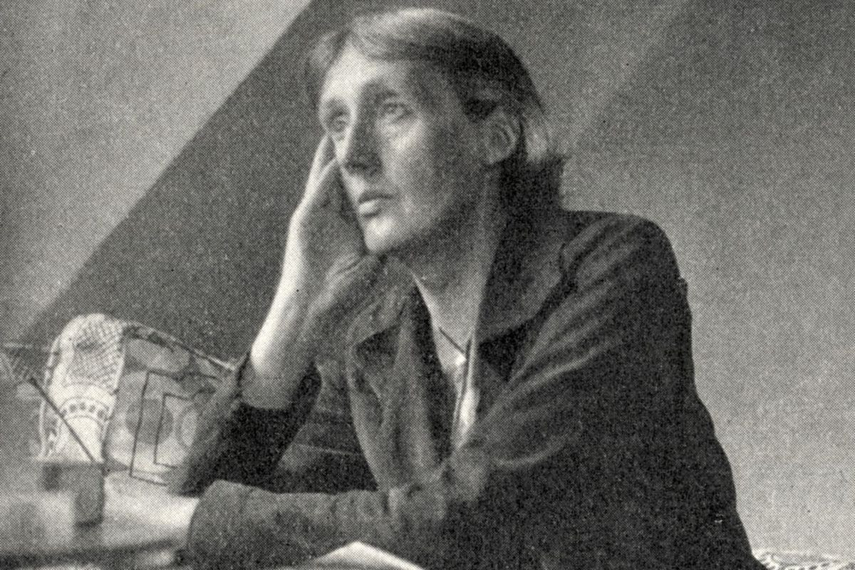 A black-and-white image of writer Virginia Woolf sitting at her desk, gazing pensively into the distance