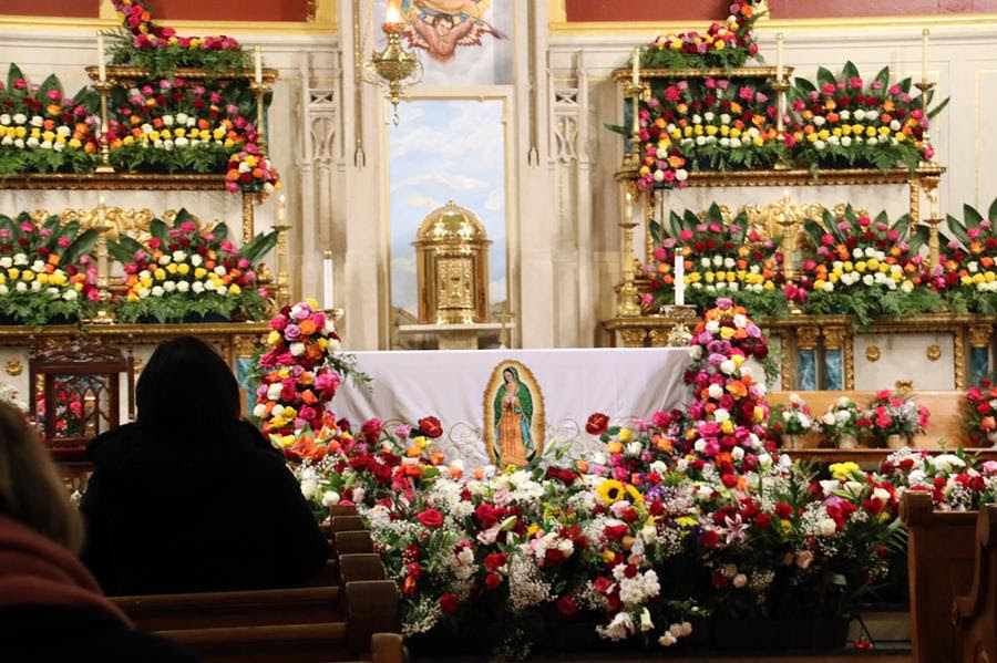 An altar in a Catholic Church with an image of Our Lady of Guadalupe. The altar is surrounded by many bouquets of colorful flowers.