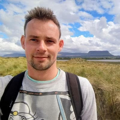 Photo showing poet Ciarán O'Rourke standing in front a bright, cloud-mottled sky, with water and mountains in the distance.