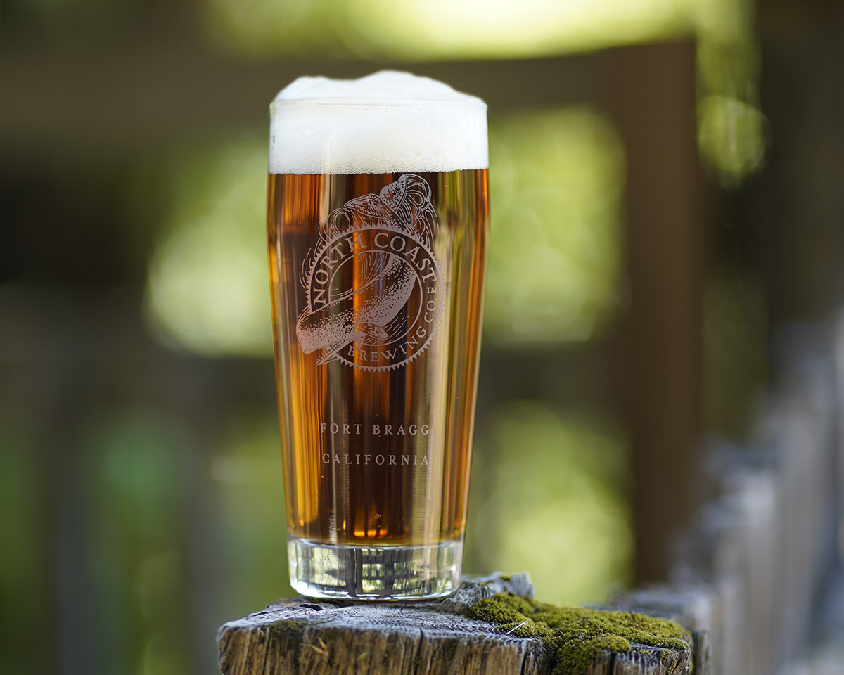 Full glass of bright amber colored beer in an outdoor setting.