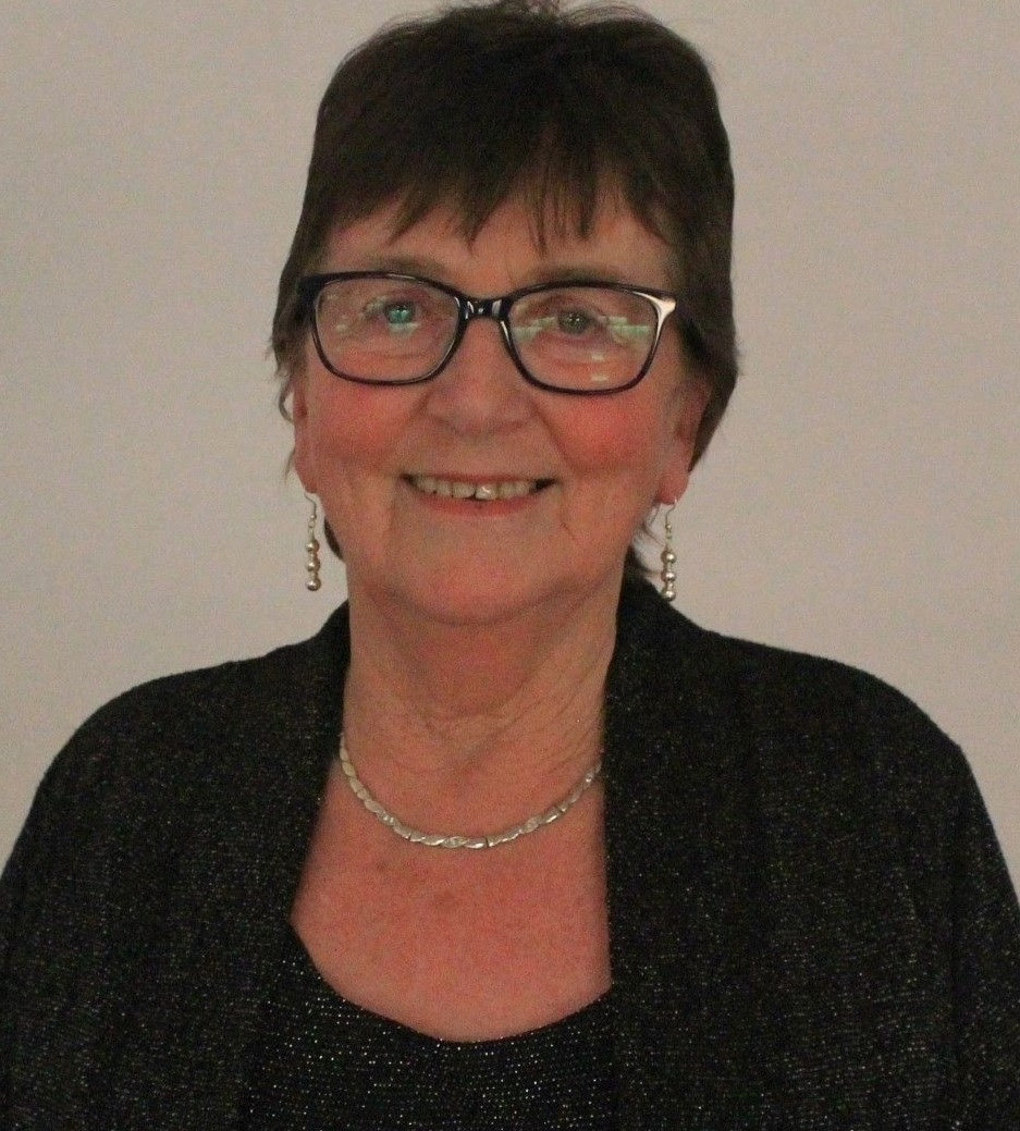 Writer Áine Ní Ghlinn stands, smiling, in front of an off-white wall, wearing glasses, silver earrings and necklace, and black clothing.