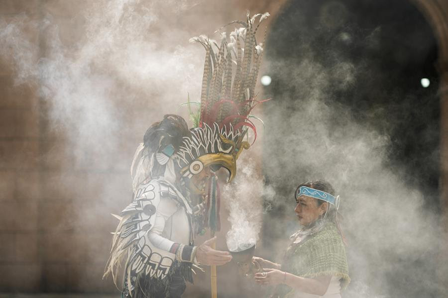 Two Mexica dancers in traditional clothing dance in a cloud of incense smoke.