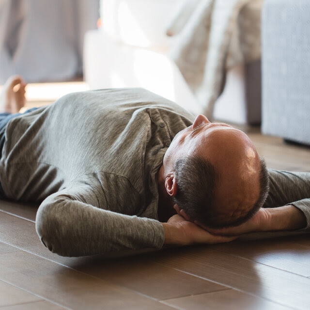 A bald man lying on a wooden floor with his hands behind his head.