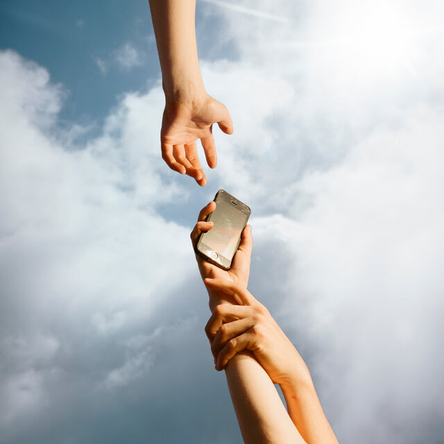Against a backdrop of clouds in the sky, a hand extends from above toward a another hand holding an iPhone, which is in turn being held back by still another hand.