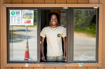 a smiling young man in a tan T-shirt with a DNR logo stands in the window of a wooden state park contact station