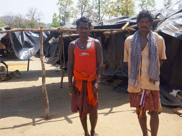 Two Khadia men stand in front of tents made from narrow trees and black tarpaulins.