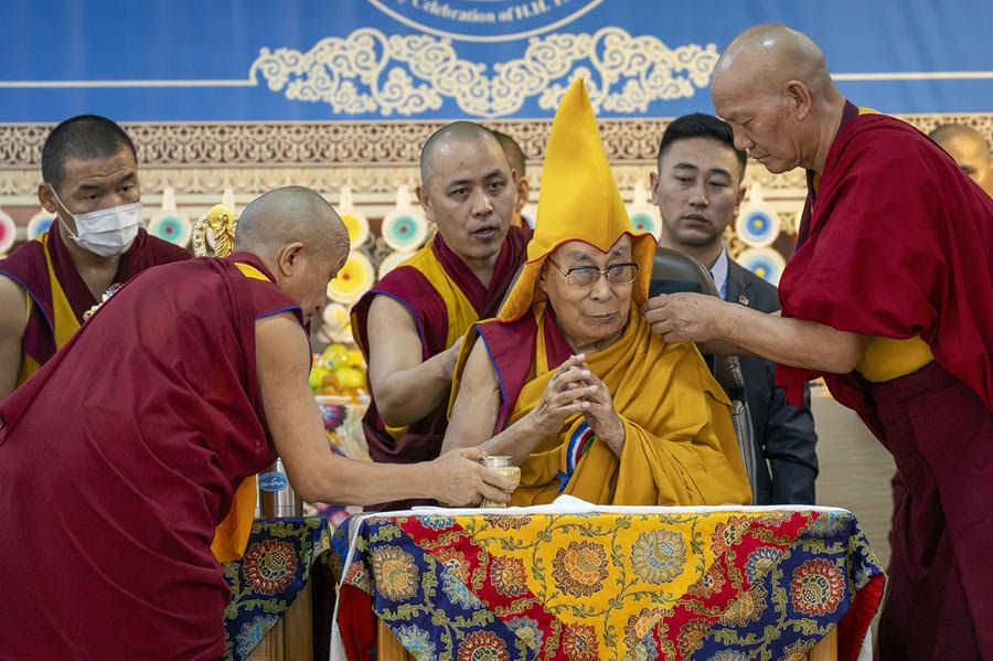 The Dalai Lama wears a ceremonial hat with monks gathered around him.