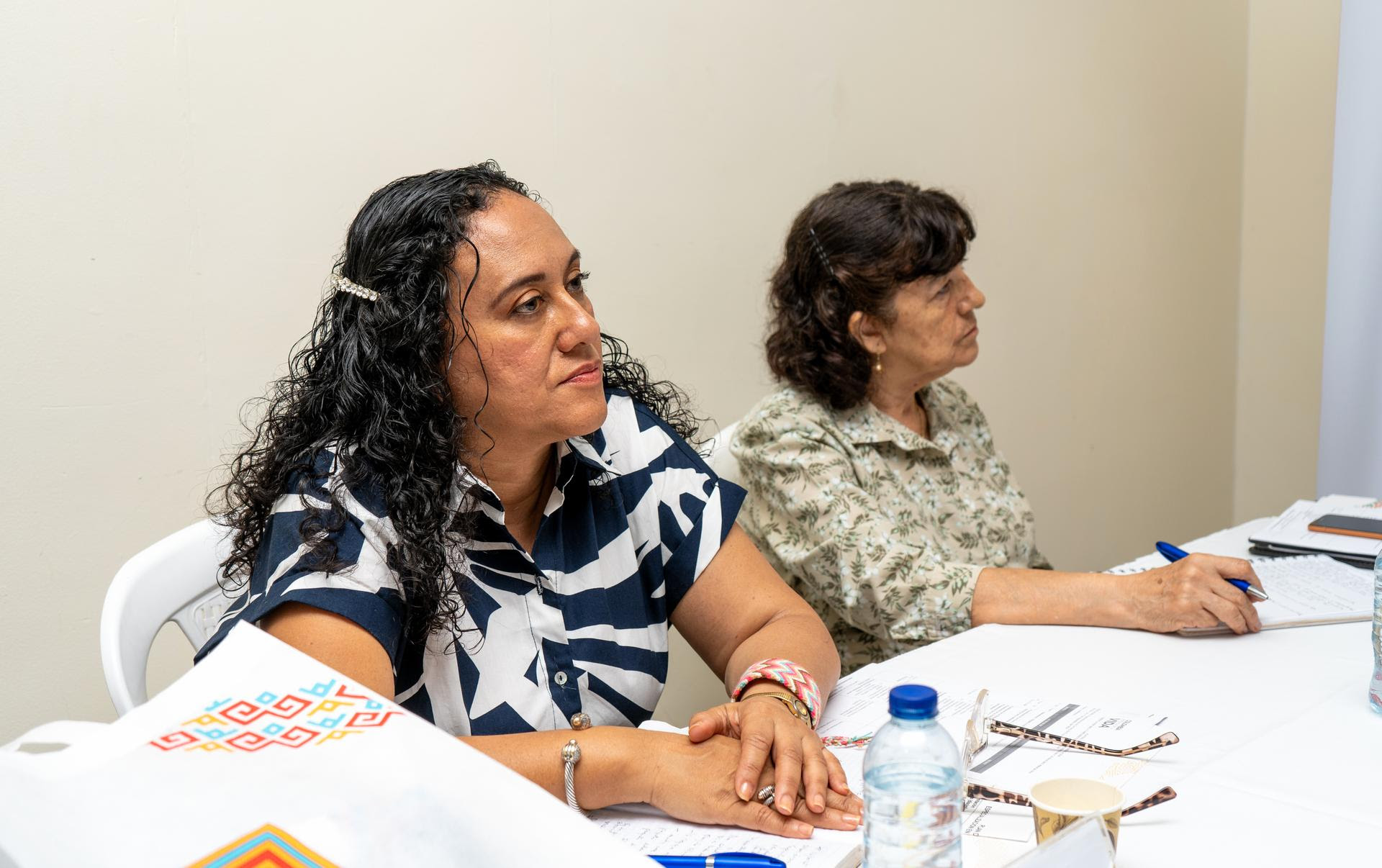 Dos mujeres están sentadas en una mesa durante una reunión o conferencia. La mujer en primer plano tiene el cabello rizado, lleva una blusa con diseño abstracto en blanco y azul, y está atenta a lo que sucede. La mujer al fondo, con el cabello recogido, viste una blusa clara con un patrón floral y está tomando notas.