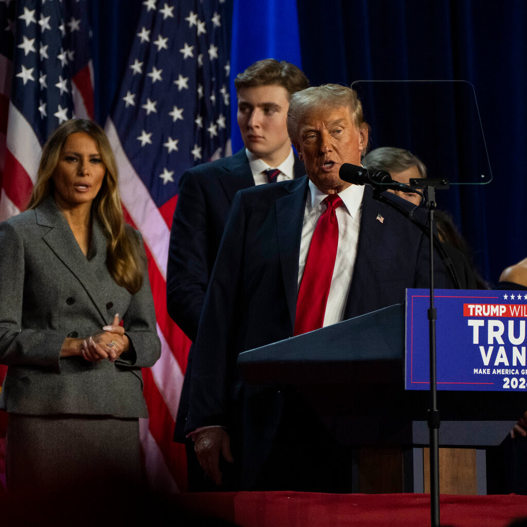 Donald Trump, wearing a dark suit and red tie, speaks behind a lectern with the words “Trump Vance” on it. 