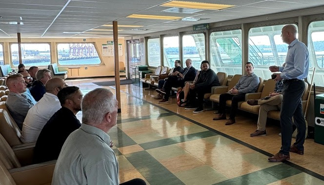 Person standing on right speaking to several people seated in the passenger cabin of a ferry