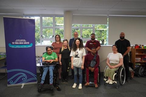 Nine people from the disabled and Deaf communities sit or stand in a row, including three wheelchair users. They are in a room with a window behind them.