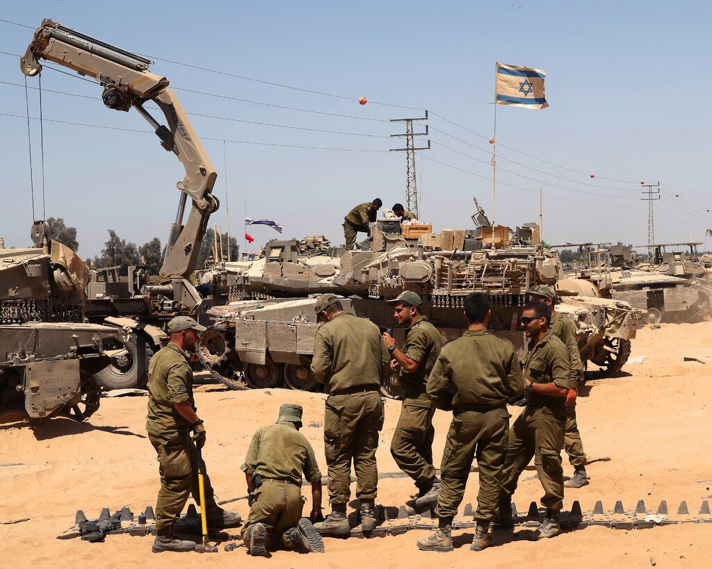 Soldiers stand around a tank tred. The ground is sandy. 