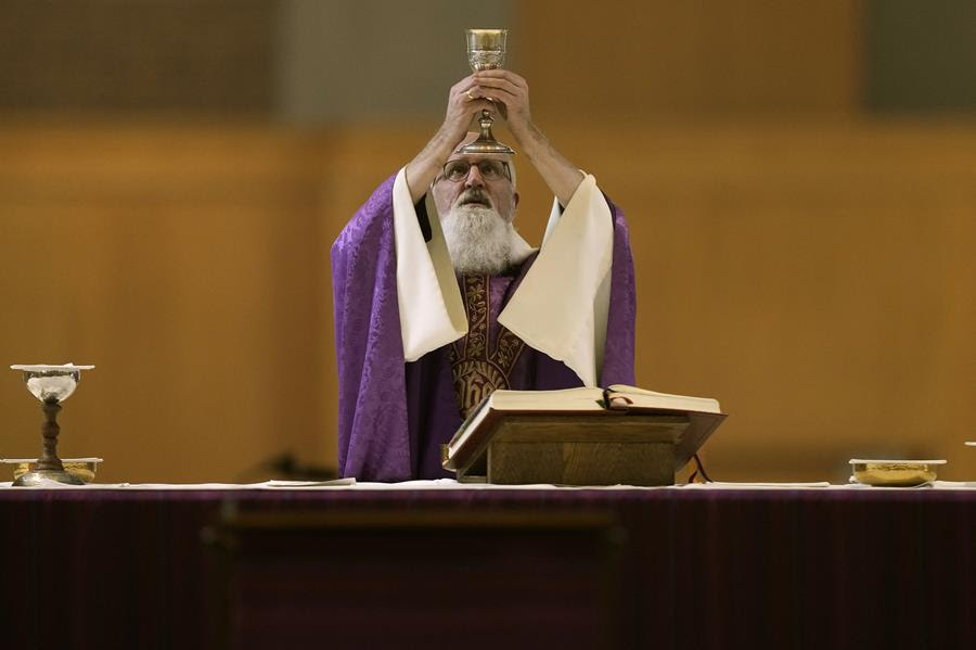 A Catholic priest wearing purple vestments stands at the altar and holds up a chalice.