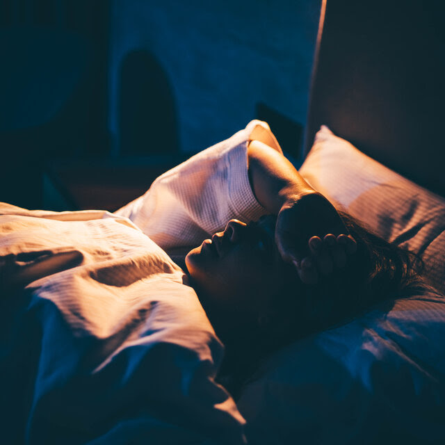 A woman lying on bed in a shadowed room with hand on forehead.