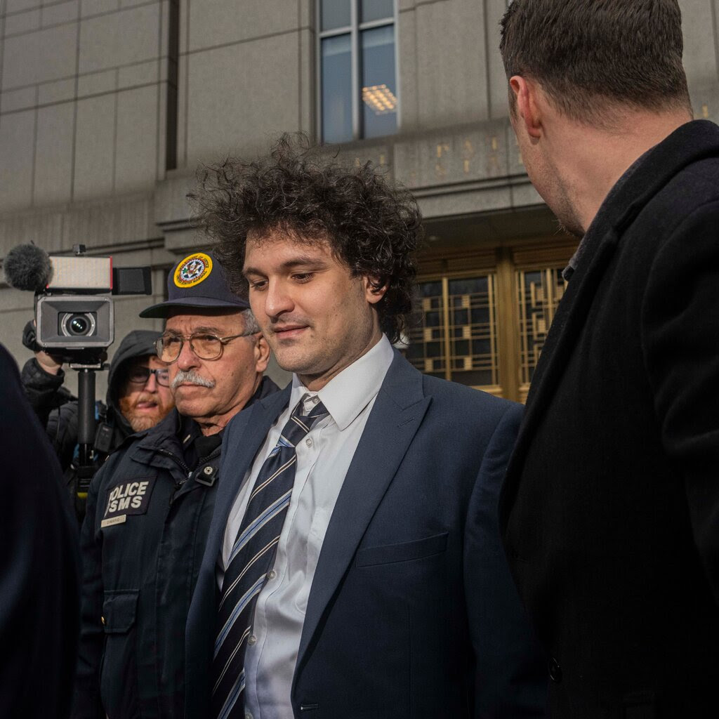 Sam Bankman-Fried, wearing a blue suit, is flanked by several people as he leaves court in New York City.