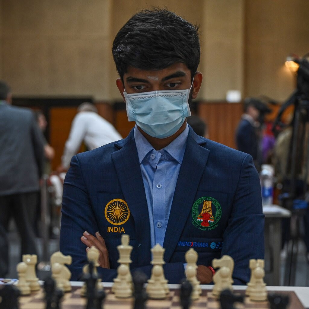 Dommaraju Gukesh, wearing a face mask, blue shirt and navy blazer, stares at a chessboard.