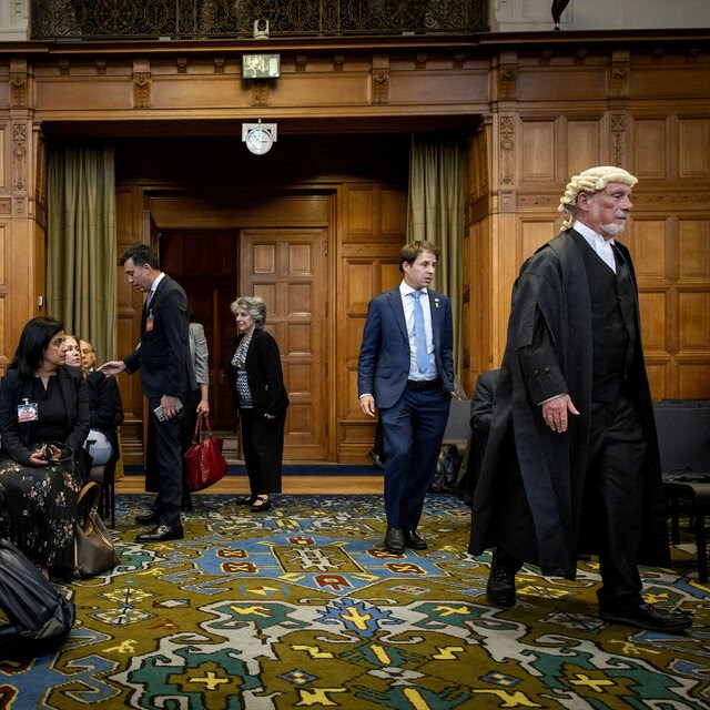 In a room with wood paneling and a multicolored carpet, several people sit and a man wearing a jurist’s black robes and white wig walks by.