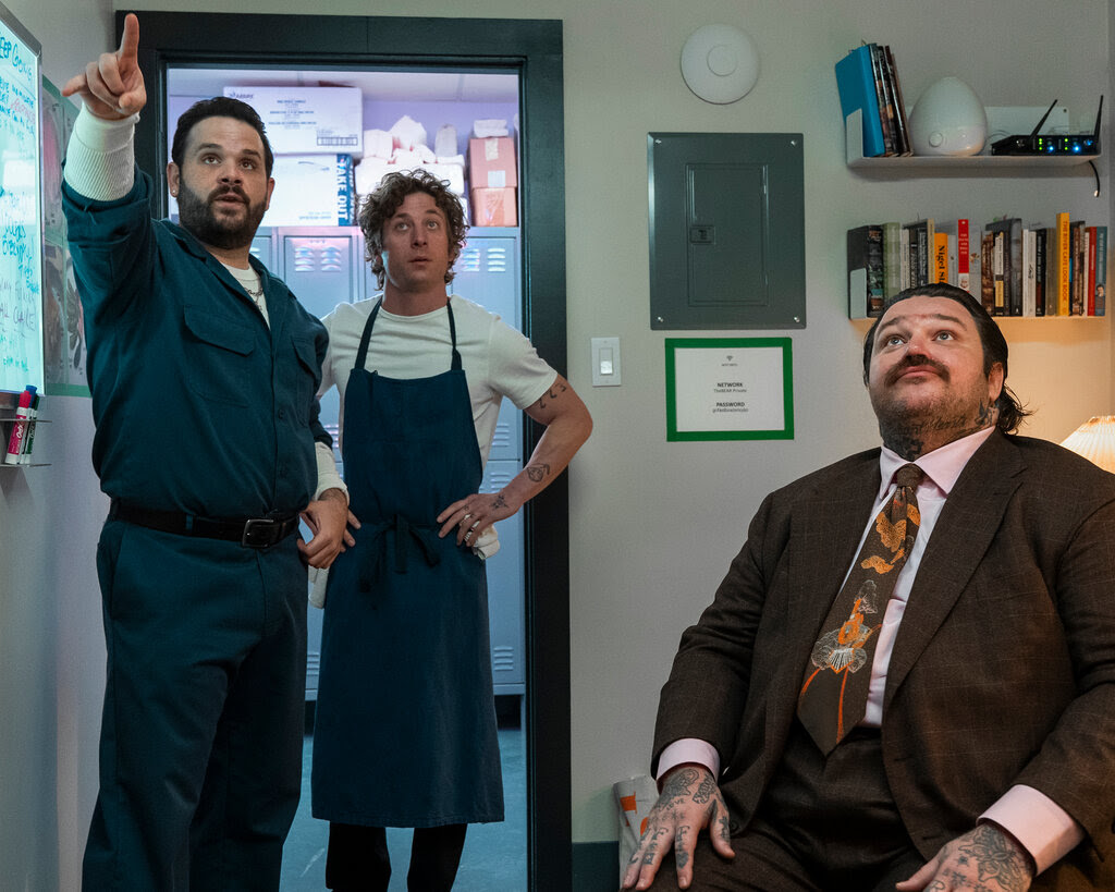 A man in coveralls points to something overhead and offscreen as another man wearing an apron and a third with hand tattoos and a brown suit look on. 