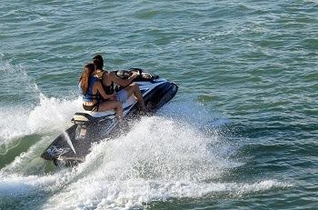 a man and woman in bathing suits and life vests ride on a Jet Ski, leaving a foamy wake as they cut through deep blue water