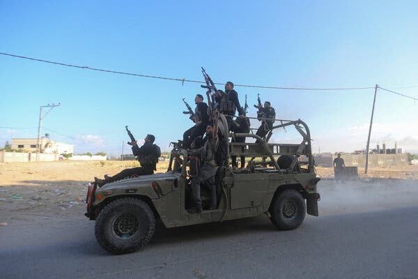 Several men holding rifles sit and ride in an olive green military vehicle driving down a dusty road.