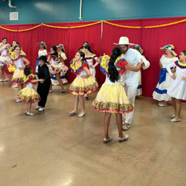 A group of dancers wearing vibrant traditional outfits performing in pairs in front of red curtains girls in colorful skirts and boys in black and white attire creating a lively and festive atmosphere