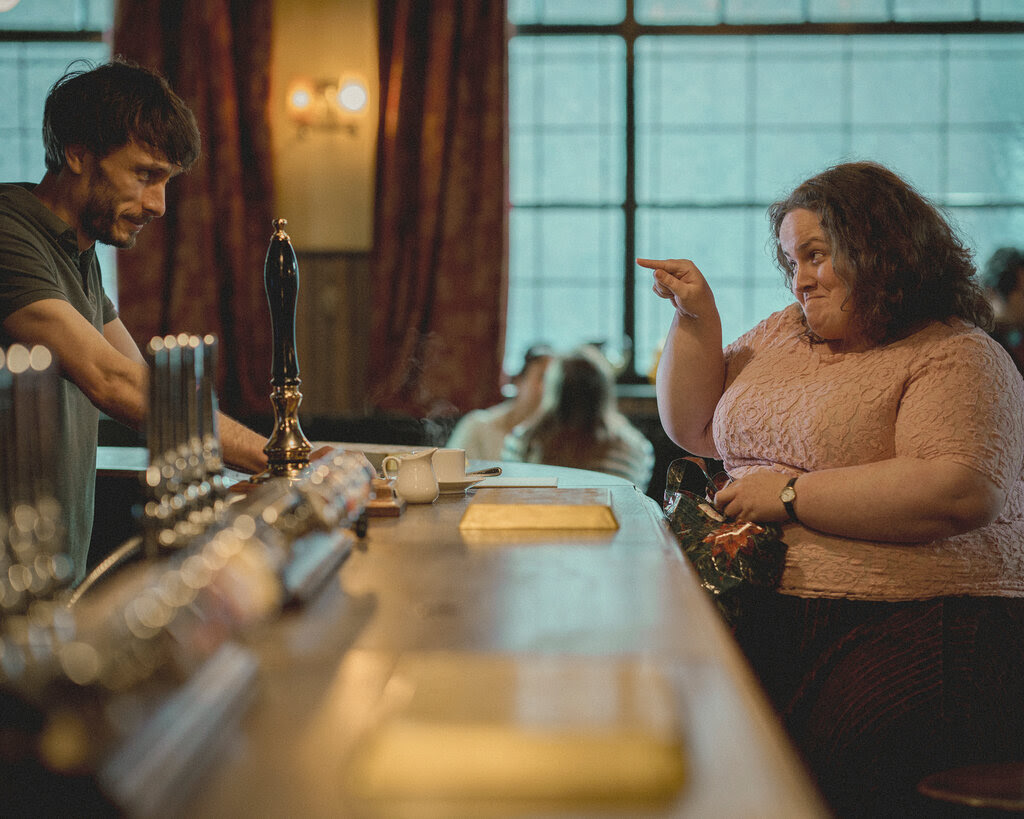 Richard Gadd stands behind the bar at a pub as a woman on a stool smiles and points at him.