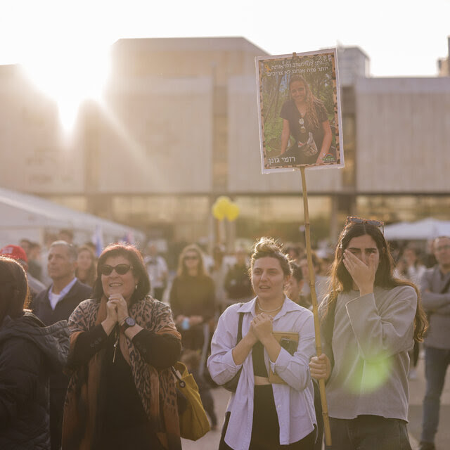 Un grupo de personas de pie en una plaza abierta, una de ellas sosteniendo una foto de una rehén israelí liberada.