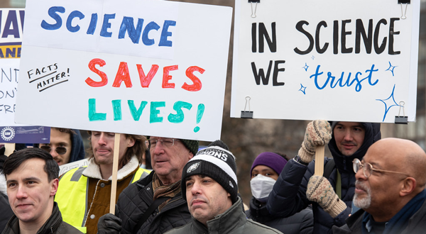 A crowd of people outdoors holding signs that read "Science Saves Lives and "In Science we Trust"