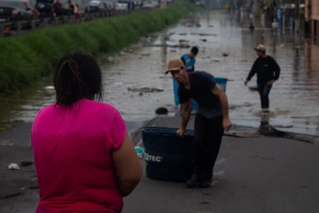 Mulher aguarda enquanto homens trazem mantimentos e cães em águas d'água às margens da BR-116