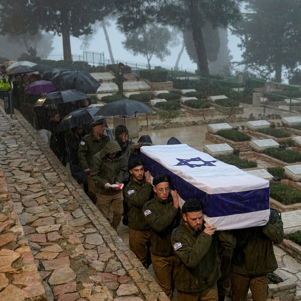 A coffin covered in an Israeli flag being carried into a tree-lined cemetery, followed by mourners holding umbrellas against the rain. 