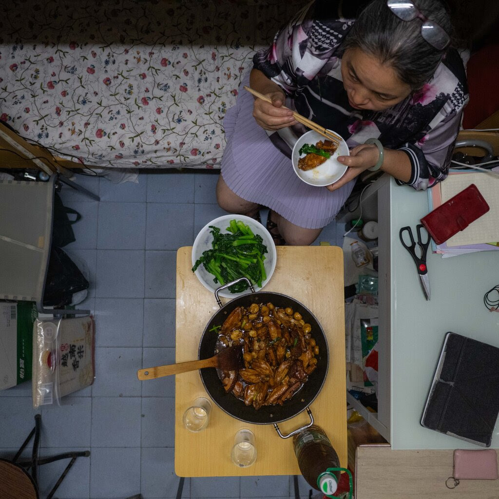 A view from above of a woman in a small, cluttered living space, eating from a bowl. In front of her is a wok with food and a wooden spatula, and a bowl of green vegetables.