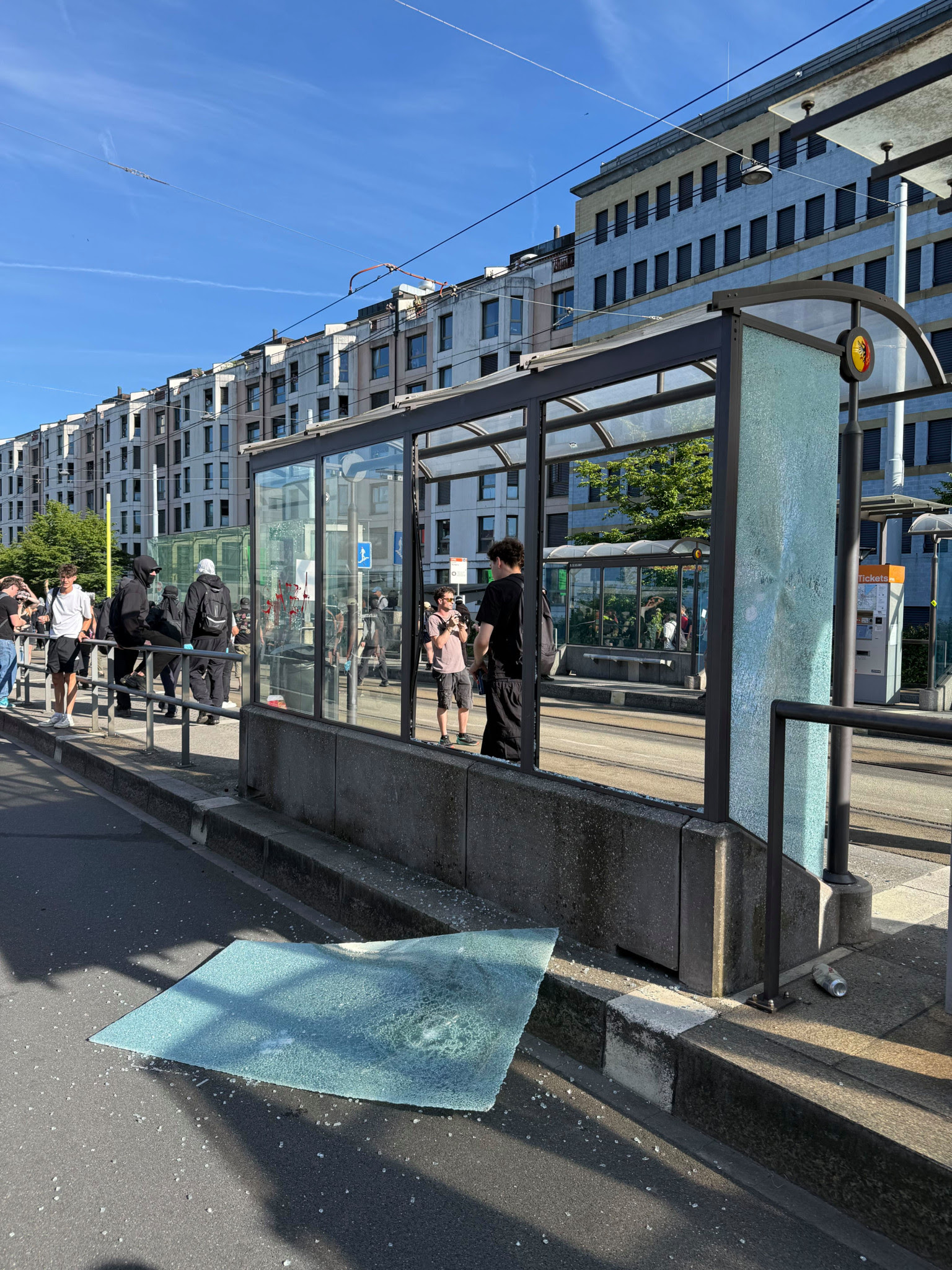 Vitre brisée d’un abri de tramway gisant sur le sol, avec des passants visibles près de l’arrêt endommagé en milieu urbain. Vitre brisée d’un abri de tramway gisant sur le sol, avec des passants visibles près de l’arrêt endommagé en milieu urbain.