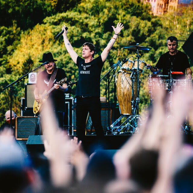 A photo of an outdoor concert, with a  man in a hat playing electric guitar, a man in a black T-shirt holding both hands in the air, and a percussionist visible behind them as fans raise their hands in the air.