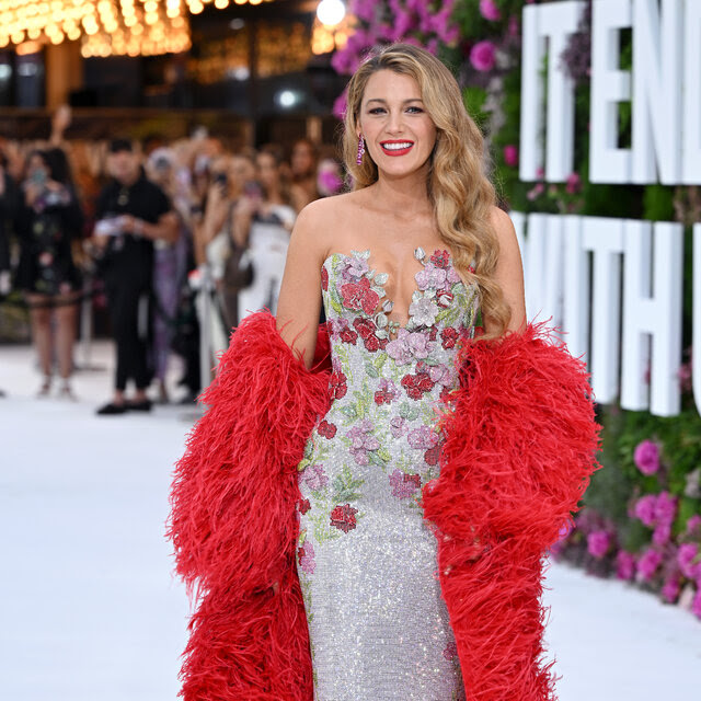 Black Lively poses on a white carpet in a long glittery dress adorned with fabric flowers and a red boa. Letters spelling out “It Ends With Us” amid real flowers are blurred in the background.