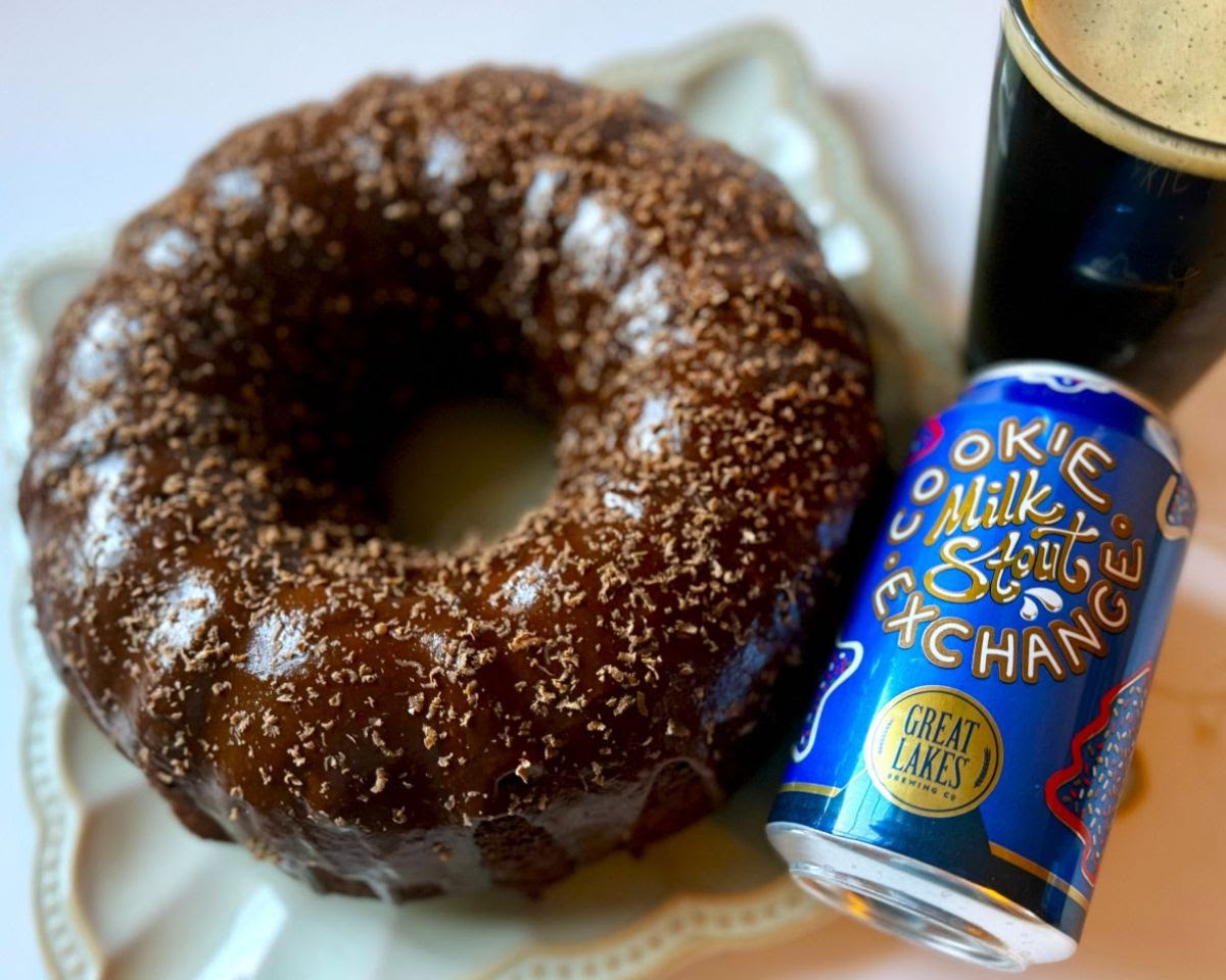 Can of Cookie Exchange Milk Stout poured into a pint glass, placed next to a dark brown chocolate bundt cake made with the beer.