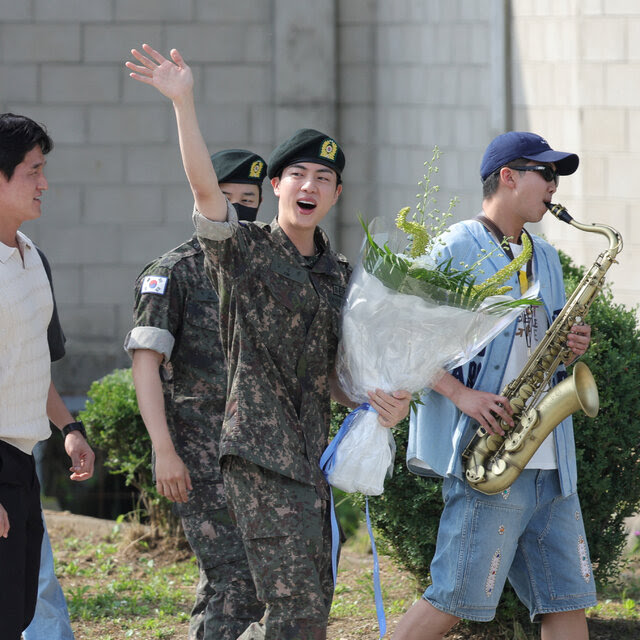 A man in military fatigues holding a large bouquet. He is surrounded by four other men, three in civilian attire. One of them is playing a saxophone.