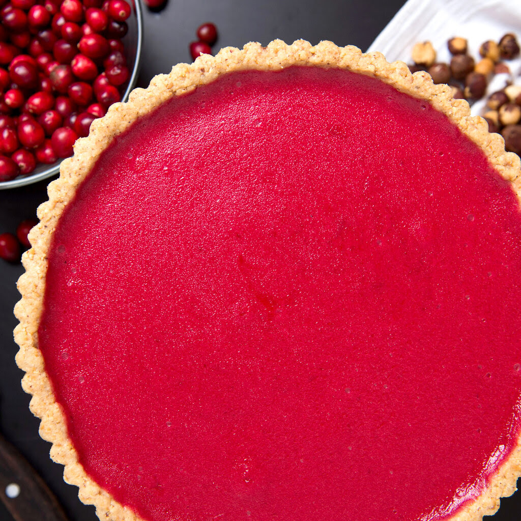 A bright red cranberry tart is photographed from overhead. A bowl of cranberries and some hazelnuts sit just above.