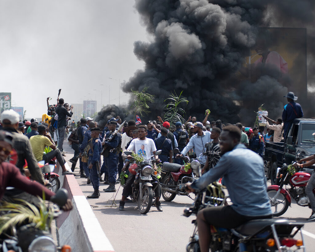 Gray smoke rises near a crowd on a street. Several people are on motorcycles.