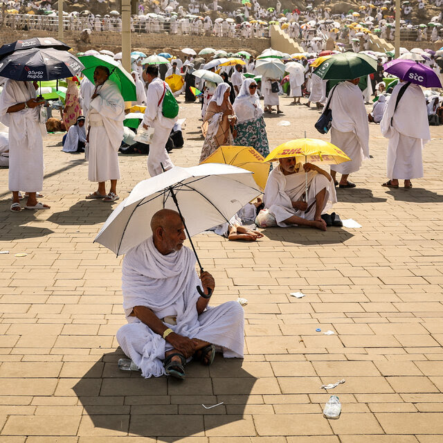 People dressed in white outdoors in a crowded area hold umbrellas. Some of people are sitting on the ground.